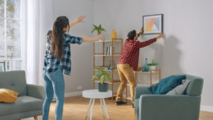 Happy Couple Hanging Picture on the Wall, Boyfriend Moves It, Girlfriend Tells Him when the Frame is Hanging Straight. Funny Moment in Young Couple's Life. Modern Stylish Apartment.