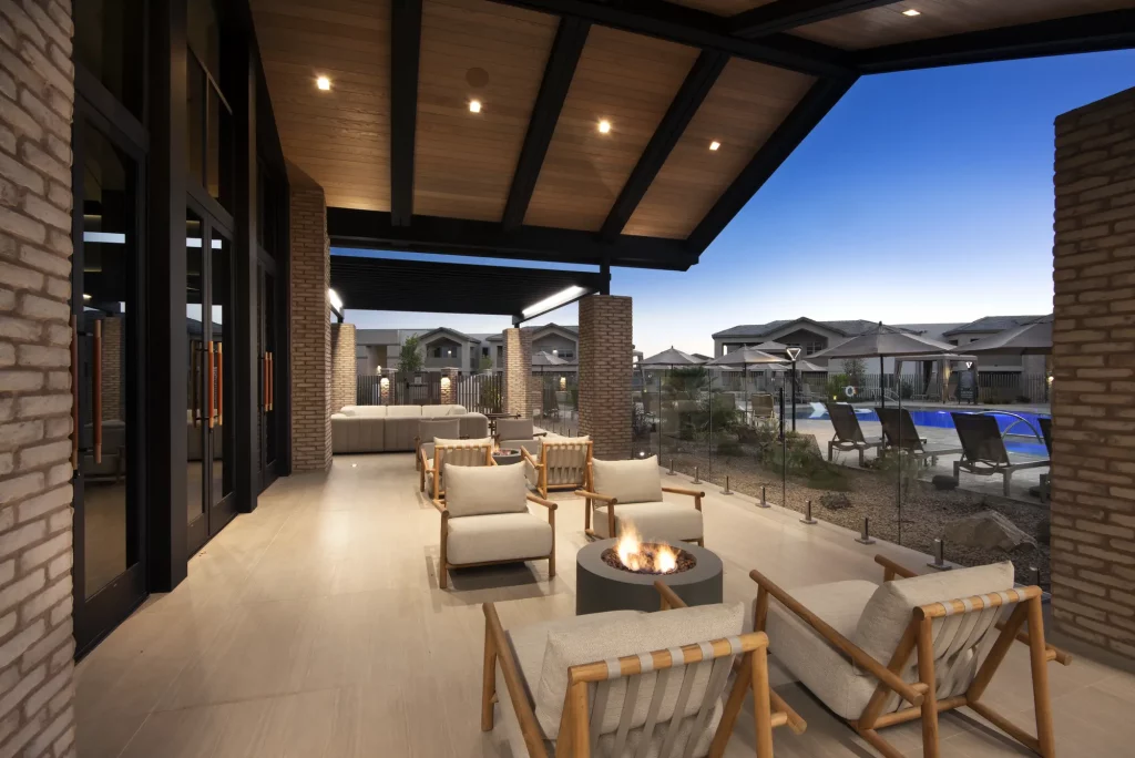 Covered outdoor lounge area at Encantada Gladden Farms featuring a lit fire pit surrounded by wood-framed chairs and neutral cushions, with a wood-beam ceiling, brick columns, glass railing, and the resort pool visible in the background at dusk.